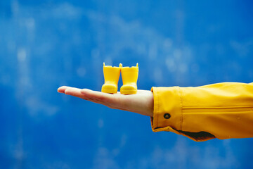 Cropped detail view of woman holding a pair of rainy boots in blue background. Horizontal close-up of hand holding little yellow galoshes isolated in blue background. Conceptual backgrounds.