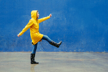Naklejka na ściany i meble Panoramic side view of woman wearing a yellow raincoat walking funny on blue wall. Full length body of woman under the rain with wellingtons and blue background. Seasonal concept winter and spring.