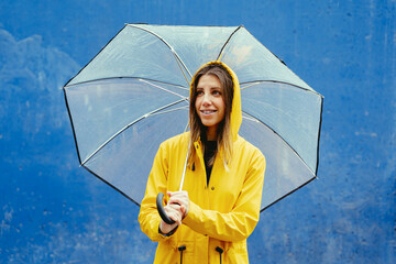 Mid waist portrait of caucasian woman in yellow waterproof clothes under rain with umbrella. Horizontal view of caucasian happy woman outdoors holding umbrella on blue background. Seasonal concept