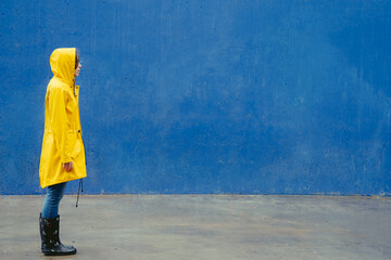 Panoramic side view of woman wearing a yellow raincoat standing on blue wall. Full length body of woman under the rain with Ukrainian yellow and blue colors. Enhanced color backgrounds.