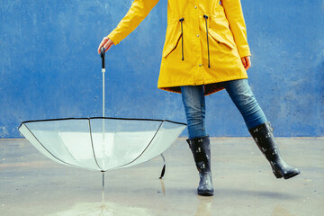 Cropped view of unrecognizable woman under rain with umbrella and rain boots. Horizontal view of woman outdoors with umbrella on blue background. Rain lifestyle concept