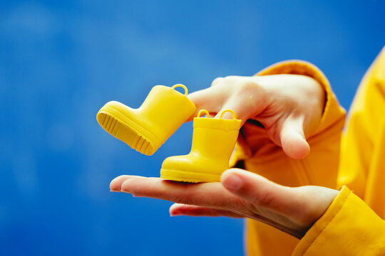 Cropped Detail Of Woman Holding A Pair Of Rainy Boots In Blue Background. Horizontal Close-up Of Hands Playing Funny Steps With Yellow Galoshes Isolated In Blue Background. Conceptual Backgrounds.