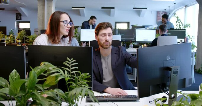 Caucasian man sitting and working a computer in office. Woman helping and giving job advice. Coworkers communicating and discussing work in coworking space. IT team cooperation.