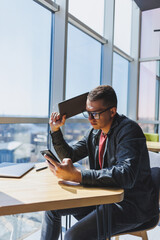 Cheerful freelancer in glasses browsing smartphone and smiling while sitting at table with laptop and notepad in cafe during daytime. Working remotely from the office.