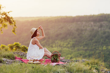 Beautiful female in trendy summer sundress and hat. She sitting on plaid on the hill and enjoying beautiful landscape.