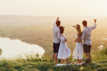 Group of friends with wine in hands enjoying summer picnic and sunset. Summer, holidays, vacation and happiness concept.