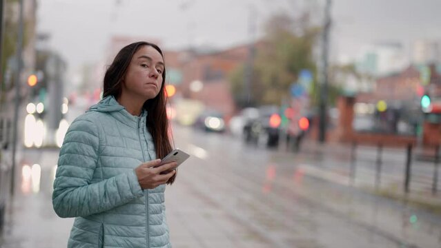 a brunette in a blue jacket, with long hair and with a phone in hand stands on a wet street on a cloudy day and looks around