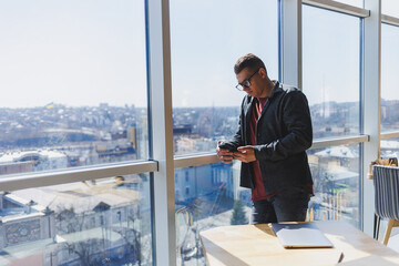 A calm confident man in a fashionable outfit and glasses stands with a smartphone in his hands near the window and looks at the city from a skyscraper in the daylight