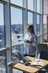 Portrait of a cheerful woman in classic glasses smiling in her free time in a cafe with coffee, positive jewish woman in a white shirt, desk with laptop, remote work