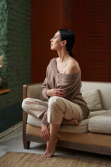 Obraz premium a beautiful woman with dark hair in loose clothes of natural colors with bare shoulders sits gracefully on a sofa near a large window