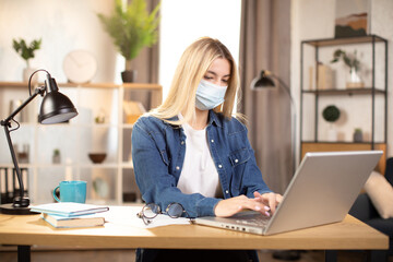 Fototapeta na wymiar Young blond Caucasian woman in face mask typing on laptop while sitting at the desk at home office. Female freelancer sitting at table and working at home during self isolation