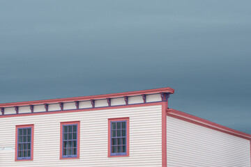 The roof section of a large white vintage wooden building with decorative pink and purple wood trim. There are three multi-pane windows on the top floor. The background is a dramatic blue cloudy sky.