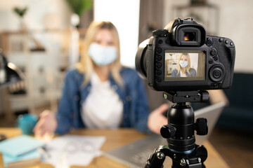 Attractive woman in protective mask using modern camera for recording video while sitting at home. Female blogger filming content for social networks. Focus on camera screen