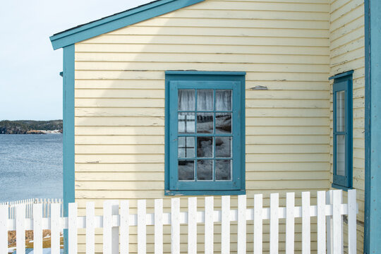 The exterior of a pale yellow wooden house with blue trim. There are two small glass pane windows in the wall. In the foreground is a white wooden picket fence. The building overlooks a blue ocean. 
