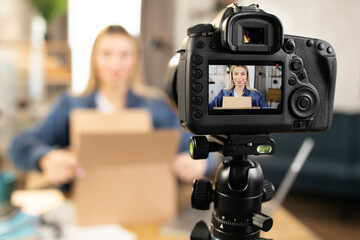 Blur background of young Caucasian blond woman unboxing parcel and recording video blog. Focus on modern digital camera. Concept of social networks technology.