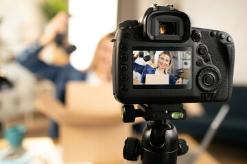 Close up of modern video camera with smiling Caucasian woman on screen. Young female influencer doing live stream while unpacking box with new headphones and joystick for games.