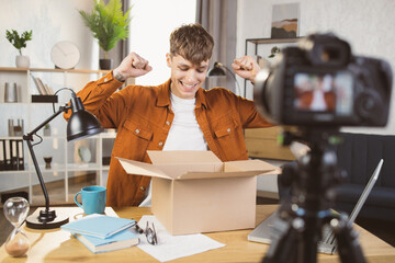 Happy young excited man in casual wear filming on digital camera unpacking process of little paper...
