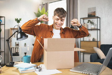 Satisfied Caucasian man sitting at desk and doing unpacking of cardboard box at home, clenches his fists in victory. Concept of people, purchases and delivery. - obrazy, fototapety, plakaty