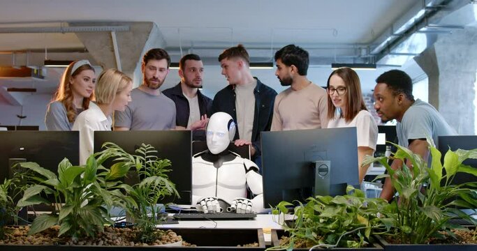 Robot sitting at table and typing on keyboard. People around applauding. Android working at computer in coworking space. Happy developers. Humanoid as IT worker. Multiethnic team cheering up.