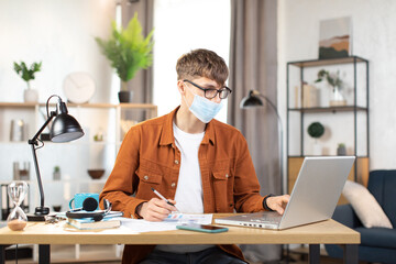 Focused young man wearing face mask, eyeglasses, sitting at table with lamp and modern laptop and making notes. Concept of technology and distance learning during pandemic.