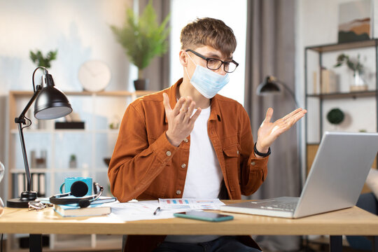 Businessman in face mask, using modern laptop for video chat at remote work. Young Caucasian guy sitting at table with laptop, lamp and papers. Technology, remote work, freelance concept.