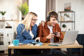 Portrait of excited focused young guy in headset, playing in video game using laptop and joystick. Pretty blond woman, sitting near man, cheering him for victory. Happy leisure time, game addiction