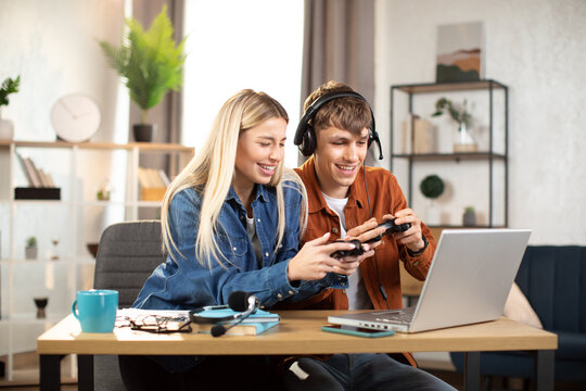 Young happy couple playing video games at home, sitting at the table with laptop and using joysticks. Happy man in headset shows new game for his lovely smiling girlfriend ar home