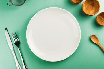 Empty white plate on the green table. Top view. Kitchen utensils beside like fork, knife, bamboo containers