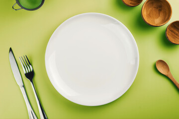 Empty white plate on the green table. Top view. Kitchen utensils beside like fork, knife, bamboo containers