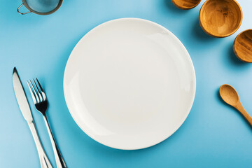 Empty white plate on the blue table. Top view. Kitchen utensils beside like fork, knife, bamboo containers
