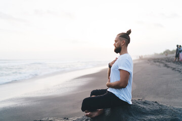 Calm Caucasian man keep praying during daytime for pranayama at seashore enjoying leisure in Indonesia, young male yogi meditate in lotus pose searching soul enlightenment and mindfulness