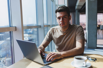 A young attractive male freelancer sits in the interior of a coffee shop and looks out the window. Handsome blogger writing ideas in laptop while sitting at wooden table in cafe with laptop