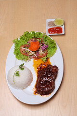 Beefsteak with rice and vegetable are placed on a white plate, on a wooden table, served with chili sauce, salt pepper and lemon