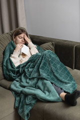 A sick girl with the flu lies on the couch covered with a blanket and holds her head