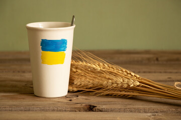 The yellow-blue flag of Ukraine is painted on a white cup with coffee on the table near the wheat...