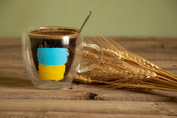 The yellow-blue flag of Ukraine is painted on a white cup with coffee on the table near the wheat...