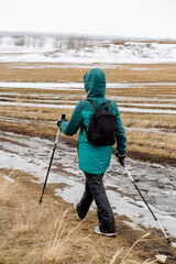 Spring hike man goes with sticks on the road, Nordic walking in nature, cardio work legs and arms, backpack black on the back, green jacket, hiker