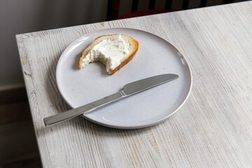 A piece of bread with curd cheese spread on it on a white ceramic plate with a knife on the beige...