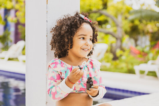 
Portrait of smiling baby girl with afro hair style and sunglasses in a colourful swimsuit outfit blowing a bubble outdoors. Afro-American and Afro-Brazilian children summer concept with copy space.