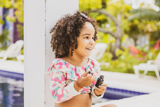 
Portrait of smiling baby girl with afro hair style and sunglasses in a colourful swimsuit outfit blowing a bubble outdoors. Afro-American and Afro-Brazilian children summer concept with copy space.