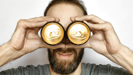 Close-up of a smiling bearded man putting two beer cans to his eyes imitating binoculars