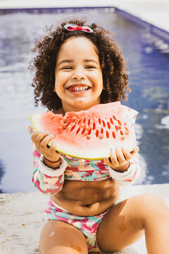 Portrait of smiling baby girl with afro hair style  in a colourful swimsuit outfit eating watermelon by the pool. Afro-American and Afro-Brazilian children summer concept with copy space.