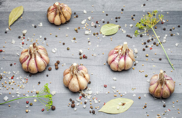 A few heads of garlic lying on a gray table top next to colorful peppercorns, bay leaf, allspice and coarse salt