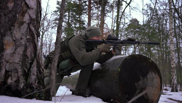 Panning shot of young concentrated sniper aiming with gun in winter forest in slow motion. Focused serious Ukrainian soldier with weapon looking away outdoors. Defense and war conflict concept