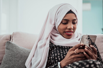 African woman using smartphone while sitting on the sofa at home wearing hijab clothes.