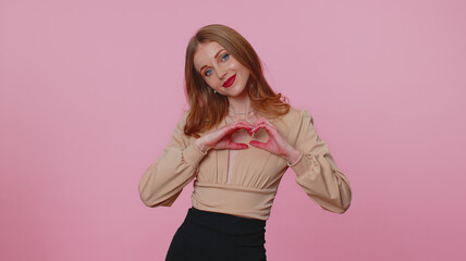 Smiling lovely businesswoman girl in beige blouse makes sign heart shape gesture demonstrates love expresses good feelings and sympathy. Young adult woman isolated alone on pink studio background
