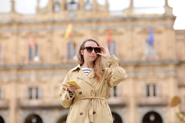 beautiful young woman with long curly hair is wearing sunglasses and using cellphone. girl out and about in the city streets during the day