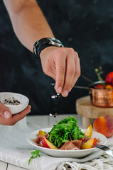 Salad bowl with grilled peach, prosciutto, goat cheese, walnuts and arugula on white background, top view, copy space. Chef salting salad