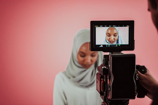 Videographer In Digital Studio Recording Video On Professional Camera By Shooting Female Muslim Woman Wearing Hijab Scarf Plastic Pink Background.