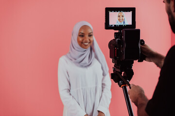 Videographer in digital studio recording video on professional camera by shooting female Muslim woman wearing hijab scarf plastic pink background.